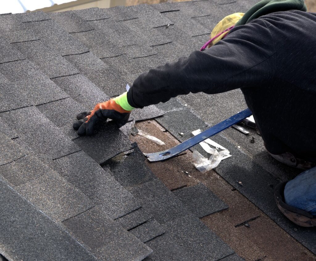 Roofer uses a blue pry bar to remove damaged shingles during the repair of a residential roof.
