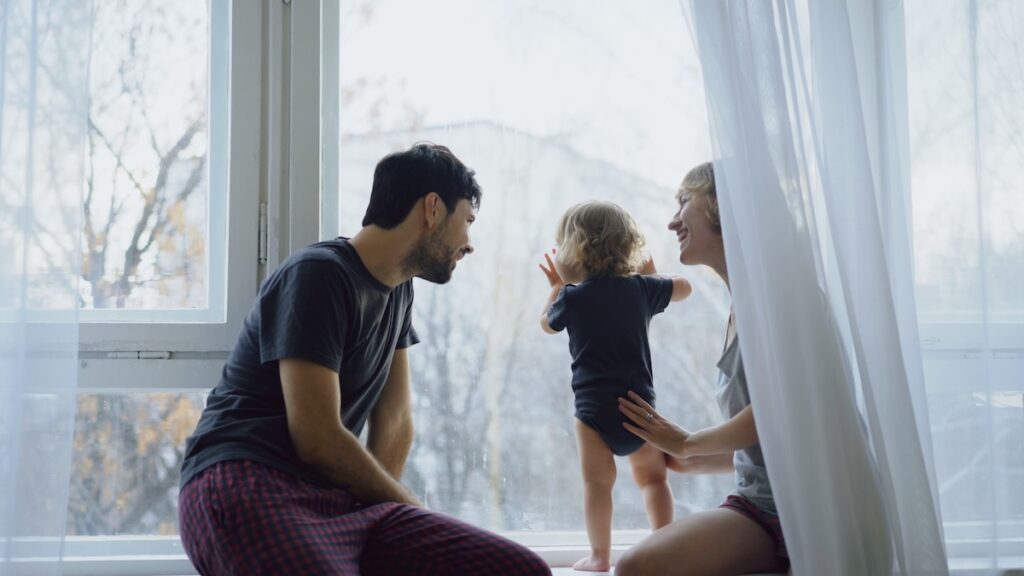 Happy family with young cute daughter sitting on windowsill playing and looking in window at home in the morning