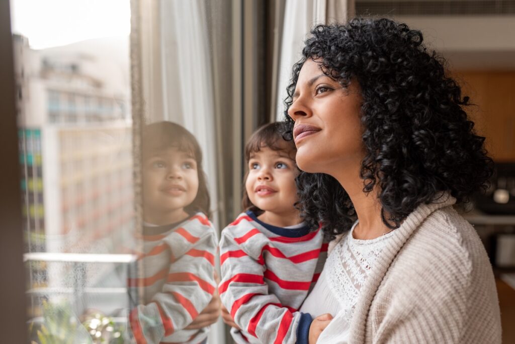 Smiling mother holding her cute little daughter in her arms while looking out through a window together at home
