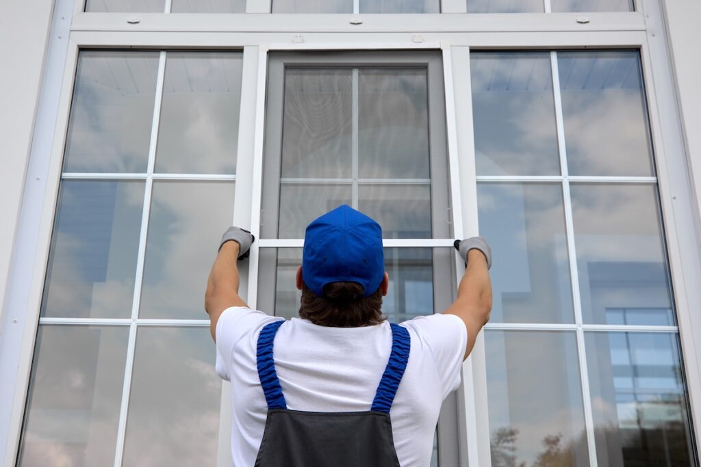 Professional craftsman outside the building installs a mosquito net on large window that lifts it up. Installation of protective net on white plastic window in summer