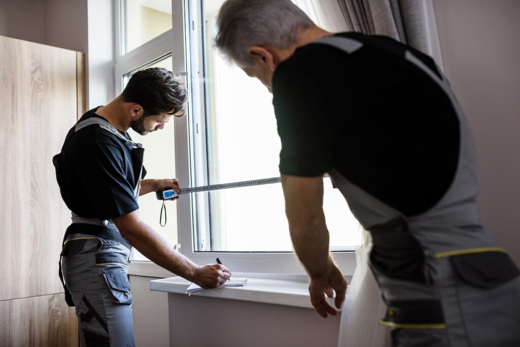 Two professional workers in uniform using tape measure while measuring window